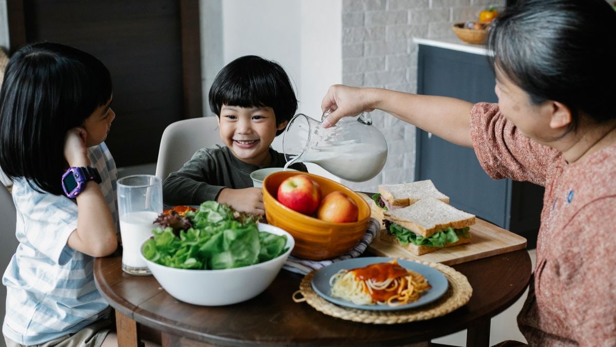 Grandmother pouring milk from jug in plate while sitting at served table with Asian boy and girl during breakfast in kitchen